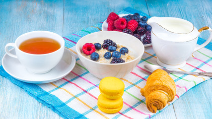 Tasty and healthy breakfast - bowl with oatmeal with raspberries, blueberries and blackberries, a cup of green tea, milk jug and croissants
