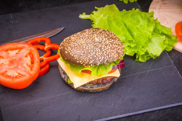 Delicious homemade dark cheese burger with vegetables on a black slate board and chopped ingredients