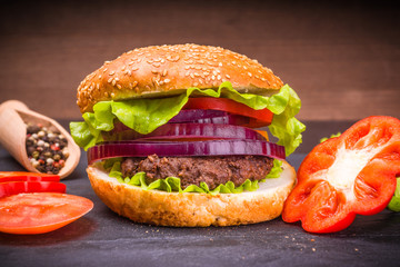 Tasty homemade burger with cutlet and vegetables on a black slate board and chopped ingredients close-up