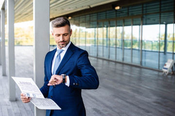 handsome businessman in formal wear holding newspaper and looking at watch