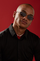 Obraz premium Close-up shot of a young man with shaved head in a black shirt and with rectangular shaped sunglasses with a black metal frame and black lenses. The photo is made on the red background.