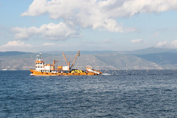 Fototapeta premium Large orange fishing boat cruising at sea with birds, mountain and clouds at background