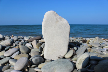 Fototapeta na wymiar Grey plain stone standing upright and small pebbles on the beach near the sea and blue sky