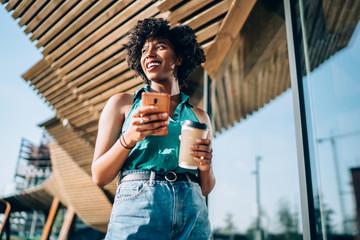 Below view of cheerful black female blogger dressed in casual wear holding coffee to go and...