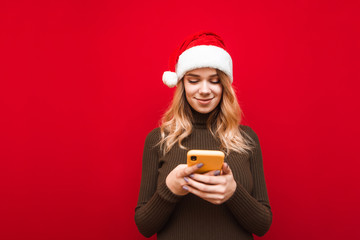 Positive lady in christmas hat uses smartphone on red background, looks at screen and smiles. Cute girl in santa claus hat uses internet on phone, isolated.