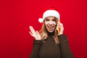 Portrait of a joyful girl in a warm sweater and Christmas hat rings on the phone against a red background, happily looks into the camera and smiles. Happy lady talking on the phone at Christmas. X-mas