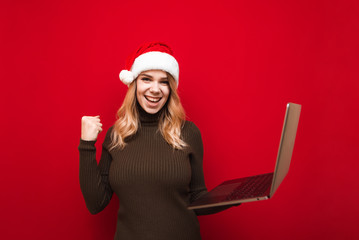 Portrait of happy girl rejoices with laptop in hand on red background, wears santa hat and warm sweater, looks into camera and smiles. Christmas girl with laptop rejoices in victory. Isolated. Xmas