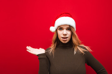 Attractive girl in a wavy hat santa stands on a red background, points her hand to the side on copy space and looks at the camera with a surprised face. New Year and Christmas cocnept.