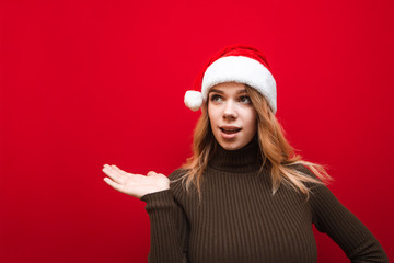 Astonished attractive girl in warm sweater on christmas hat santa claus looks and points with her hand to the empty space, isolated. Emotional girl holding copyspace in her hand on red background.
