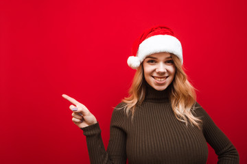 Happy girl in santa claus hat wearing hat stands on red background, wears warm sweater, shows thumbs up at copy space, looks into camera and smiles. New Year and Christmas concept