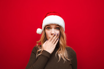 Closeup portrait of shocked girl in warm sweater and santa hat on red background, looks into camera and closes mouth with surprise by hand. Isolated. New Year and Christmas concept.