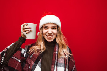 Christmas portrait of happy girl in plaid and santa hat on red background, holds big cup in hand with warm drink, looks into camera and smiles. Joyful girl drinking tea at Christmas, isolated.