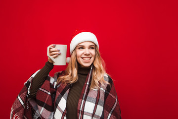 Attractive girl in plaid and christmas hat and plaid stands on red background with cup of hot drink in her hand, looks away and smiles. Isolated christmas portrait of girl with cup of tea. Xmas