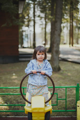 brunette girl 2 years old in a dress and jacket is standing on a yellow swing on the playground 