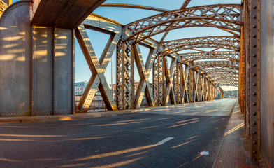 Asphalt road under the steel construction of a bridge in the city on a sunny day. Evening urban scene with the sunbeam in the tunnel. City life, transport and traffic concept.	