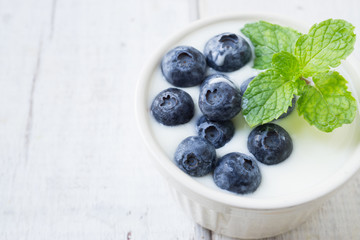 Close up white yogurt with blueberries and mint leaf topping on white cup on white wooden table.