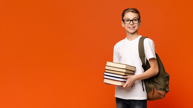 Positive teen boy in eyeglasses with backpack holding heap of books