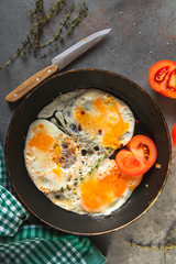 fried eggs with tomato and thyme and spices on a black skillet on a gray background, knife, towel in a green cage,