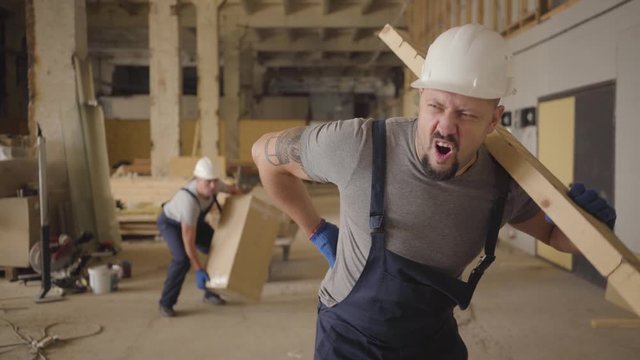 Adult Caucasian Man In White Helmet And Blue Uniform Walking At The Construction Site With A Wooden Plank. Builder Stopping Because Of Sharp Pain In Back, Another Worker Running Up To Help.