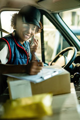 Happy black deliverer talking on the phone while filling paperwork in a van.