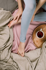 Cozy flatlay of woman's legs in blue leggins in bed with cup of tea, selective focus