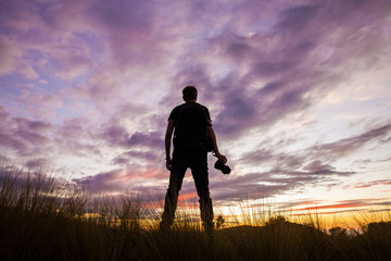 Rear view of a young man standing with his photo camera and watching a colourful sunset with purple...