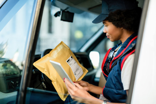 African American courier checking address on a package while using touchpad in delivery van.