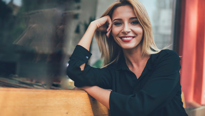 Close up portrait of pretty Caucasian woman with blonde hair taking rest on city bench and looking at camera with cute sincerely smile on face, positive hipster girl feeling good during free day - obrazy, fototapety, plakaty