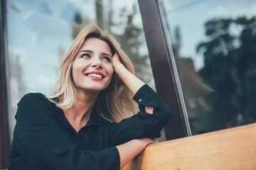 Tuinposter Positive cheerful hipster girl with perfect blonde hair taking rest on bench in city and enjoying time for dreaming outdoors, happy female with cute candid smile looking away during leisure © BullRun