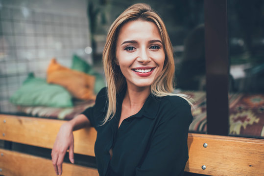 Close up portrait of attractive Caucasian millennial girl in casual wear sitting on city bench and smiling at camera, happy carefree hipster woman with perfect white teeth taking rest outdoors