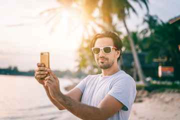 Stylish sexy young brunette man with a beard in sunglasses on a tropical beach with a mobile phone in his hands at sunset