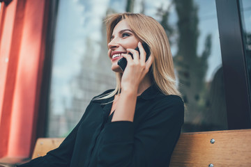 Cheerful Caucasian hipster girl with blonde hair smiling during positive smartphone conversation with friend, happy woman using 4g roaming internet connection for phoning via cellphone application