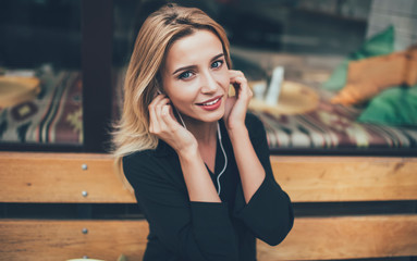 Close up portrait of young female spending time for listening audio book via electronic headphones enjoying leisure outdoors, attractive woman in black shirt looking at camera enjoying music song