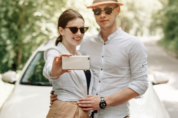 Young Happy Couple Dressed Alike in White T-shirt Holding Smartphone Near the Car, Weekend Outside the City, Holidays and Road Trip Concept