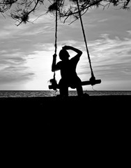 Silhoutte image of young boy drinking water on the swing,kid having fun by playing wooden swing on the beach in sunset,black and white style