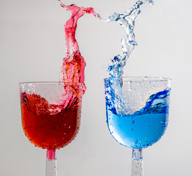 Stop Motion Image Of Two Wine Glasses With Red And Blue Liqid Splashing Into The Shape Of Woman And Man Dancing Together With Heart Shape In Negative Space On White Backround.