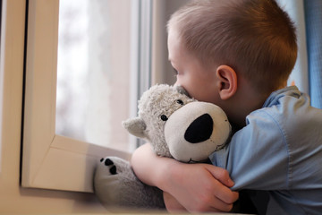 Sad boy at the window and bear toy, loneliness and depression