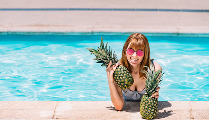 Girl with sunglasses leaning on the edge of the pool with pineapples and sticking out her tongue