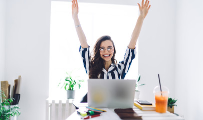 Happy young female sitting at desk with laptop and raising hands up