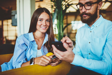 Portrait of successful female teenager smiling at camera while male friend sitting near and checking notification on telephone device using 4G internet, hipster guy sending sms message on smartphone