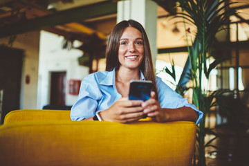 Portrait of young positive female teenager sitting indoors and holding modern mobile phone, caucasian smiling woman looking at camera during installing multimedia application on cellular device