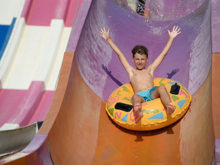 Boy on floater sliding down slide in waterpark. His hands are wide open.