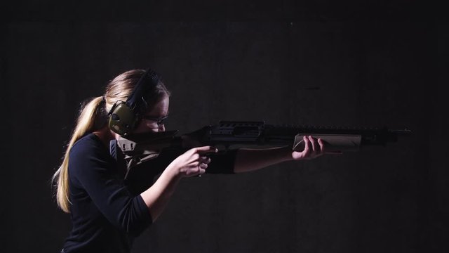 Young Blonde Female with Ear and Eye Protection on and a Shot Gun in Hand Aiming and Firing in a Shooting Range Indoors
