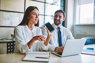 Fototapeta premium Young attractive positive female person holding mobile phone in hand and explaining to colleague during sitting at wooden table in office.Charming woman graphic designer working with creative male