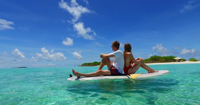 Couple sitting on surfboard, floating in ocean, boy has white t-shirt and sunglasses, young girl has red swimsuit on
