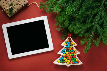 Christmas composition. A plate in the shape of a Christmas tree filled with colorful candies. Fir branches, Christmas decorations and gadget on a red background. Flat lay, top view, copy space.