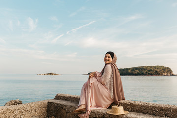 portrait of young European Muslim women with hijab sitting on the beach with hat in her hand. She is happy and relaxed. Sea is in the background. She is looking at the camera.
