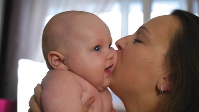 Mother and Baby. Happy young mother hugs baby and kisses on chubby cheeks at home. Close up shot.