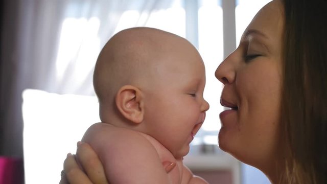 Mother and Baby. Happy young mother hugs baby and kisses on chubby cheeks at home. Close up shot.
