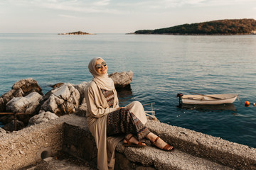 portrait of young European Muslim women with hijab sitting on the stone beach with sea in the background. She is happy and relaxed.